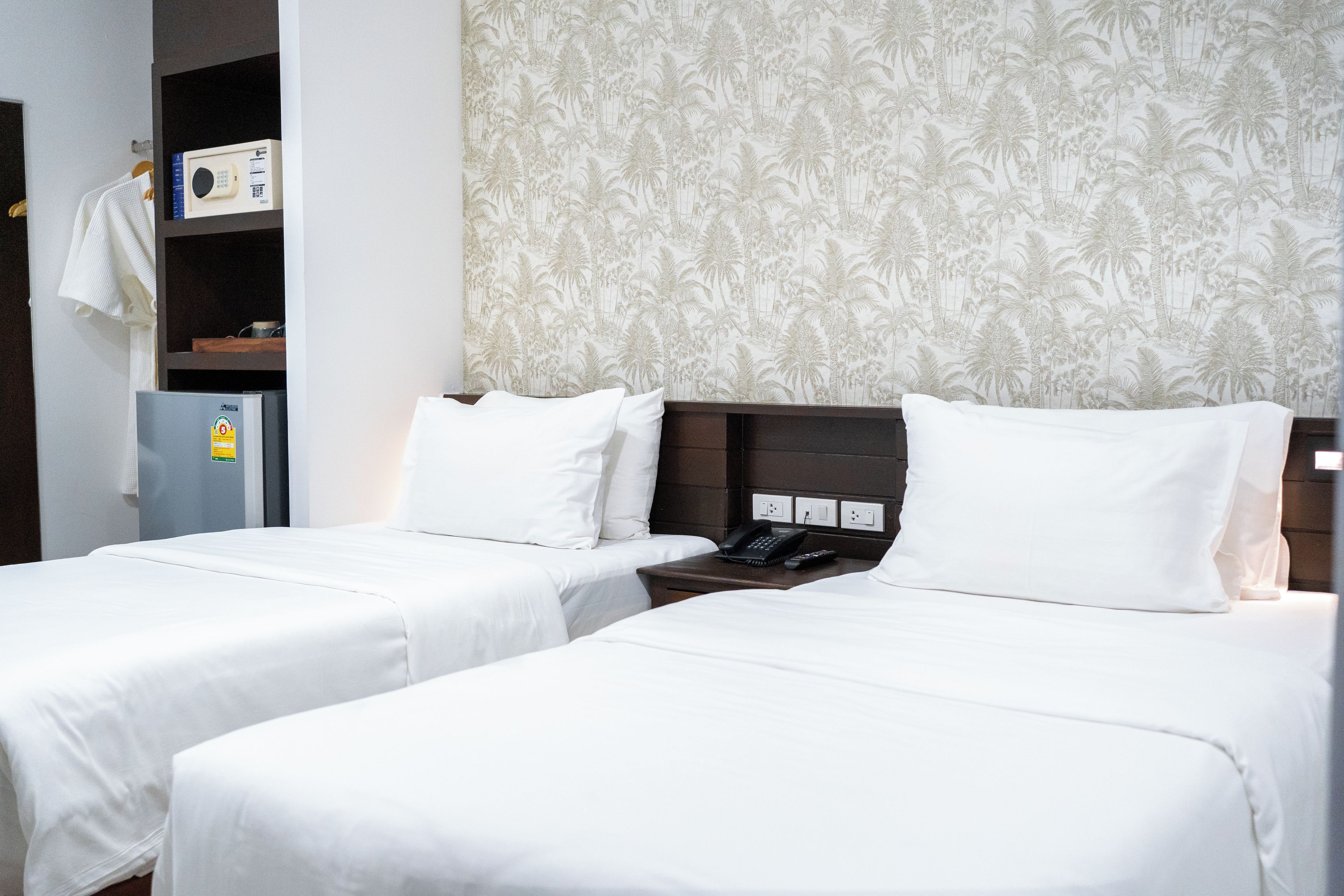 A hotel room with two beds, a wardrobe, a desk with a telephone, and a wall with a floral pattern.