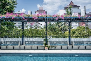 Ein Poolbereich mit einer Pergola, die mit blühenden Pflanzen bewachsen ist, Betonbänken und einem klaren, blauen Pool.