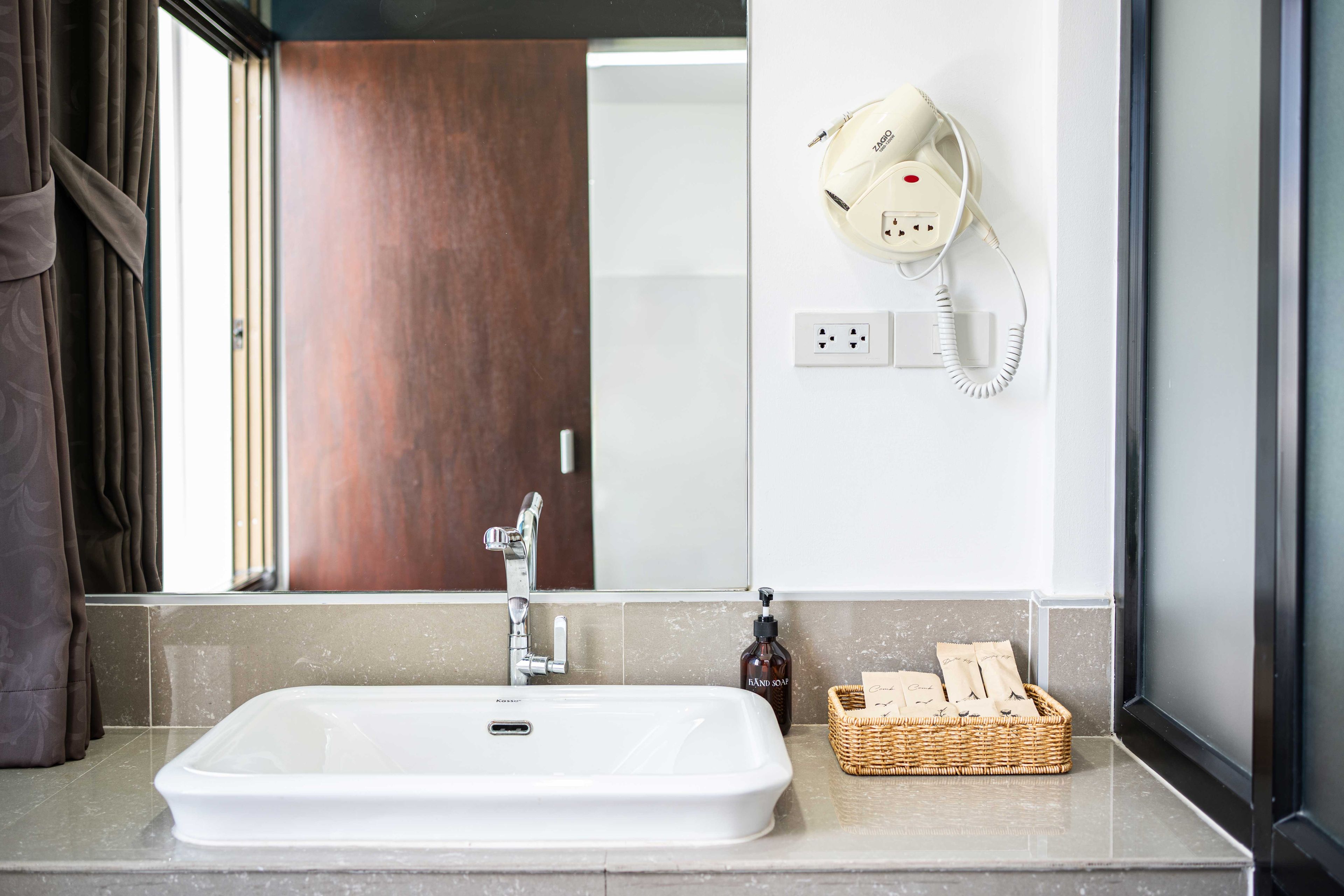 A bathroom with a white sink, a mirror, a hairdryer on the wall, a bottle of hand soap, and a basket with towels.