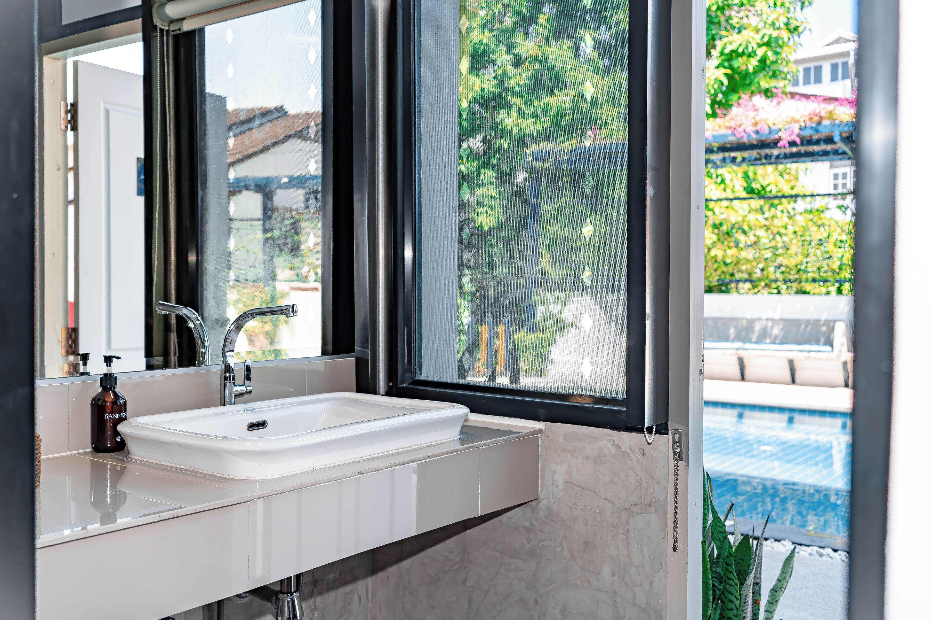 A bathroom with a white sink, a mirror, and a window with a view of a swimming pool and greenery.