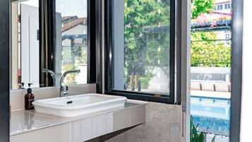 A bathroom with a white sink, a mirror, and a window with a view of a swimming pool and greenery.