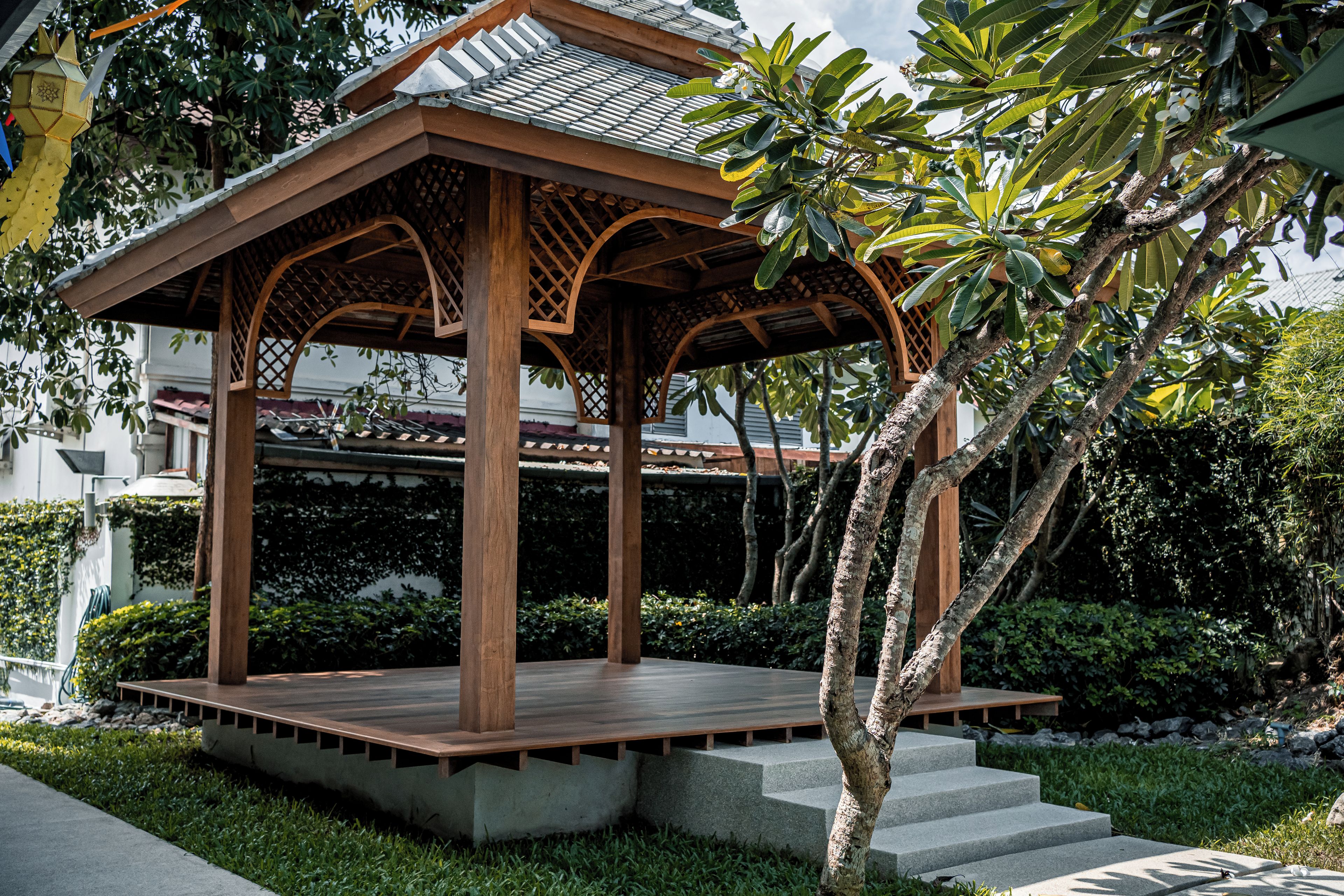 A wooden gazebo with a tiled roof, surrounded by greenery and a staircase leading up to it.