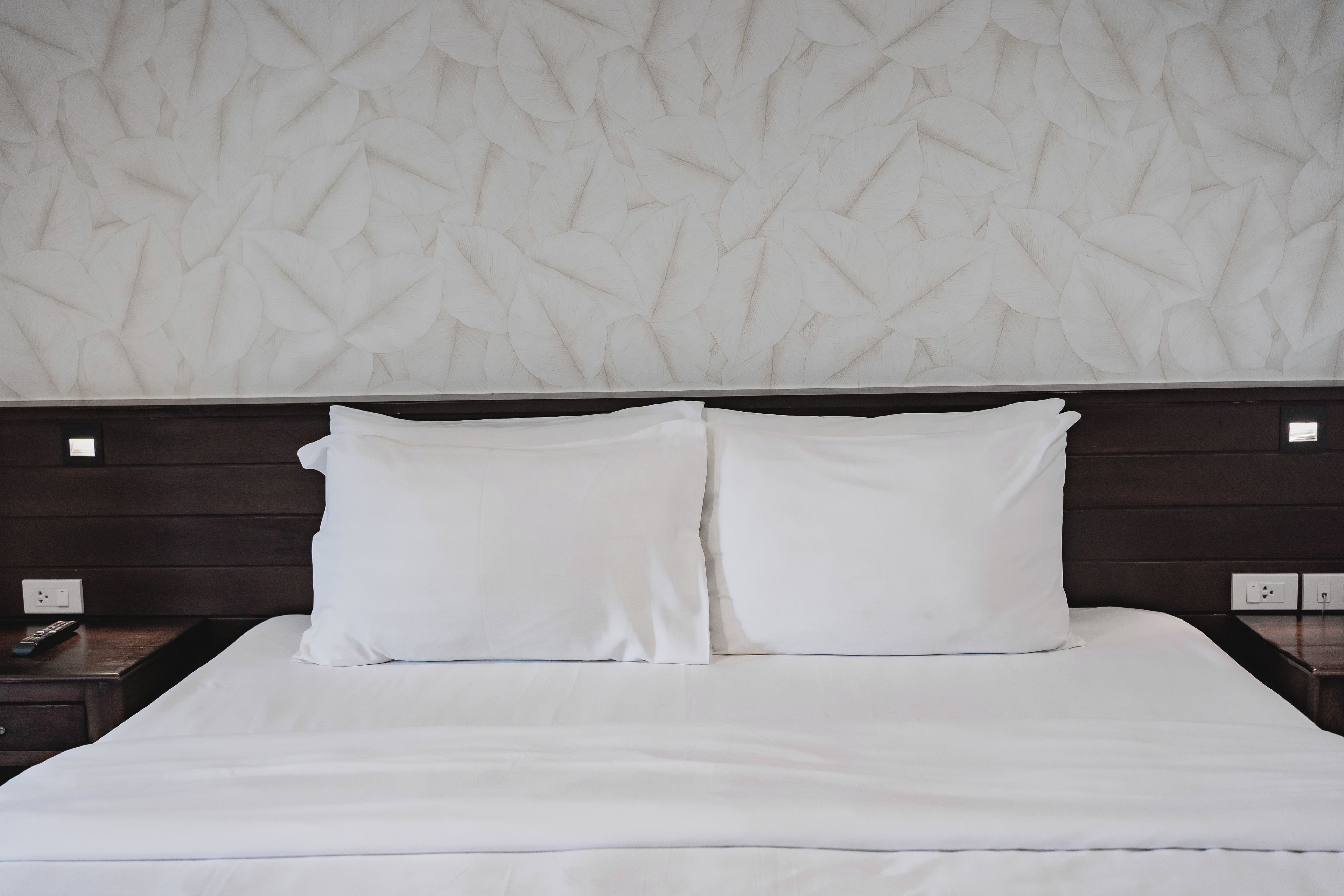 A hotel room with a large bed, two pillows, a wooden headboard, and a wall with a leaf pattern.