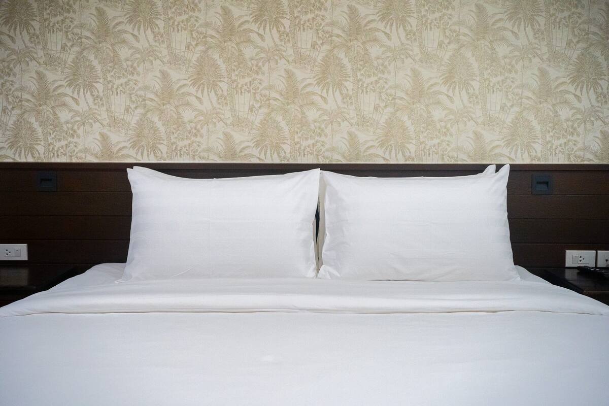 Een netjes opgemaakt bed met witte lakens en een hoofdbord tegen een muur met een palmboommotief.