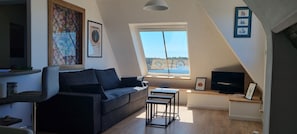 A modern loft with a dark gray sofa, a small wooden table, a TV, and a large window offering a view of the sea.