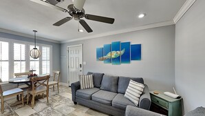 Ein Wohnzimmer mit grauem Sofa, einem Esstisch mit Stühlen, einem Deckenventilator und einer Wand mit einem Gemälde von einem Seepferdchen.