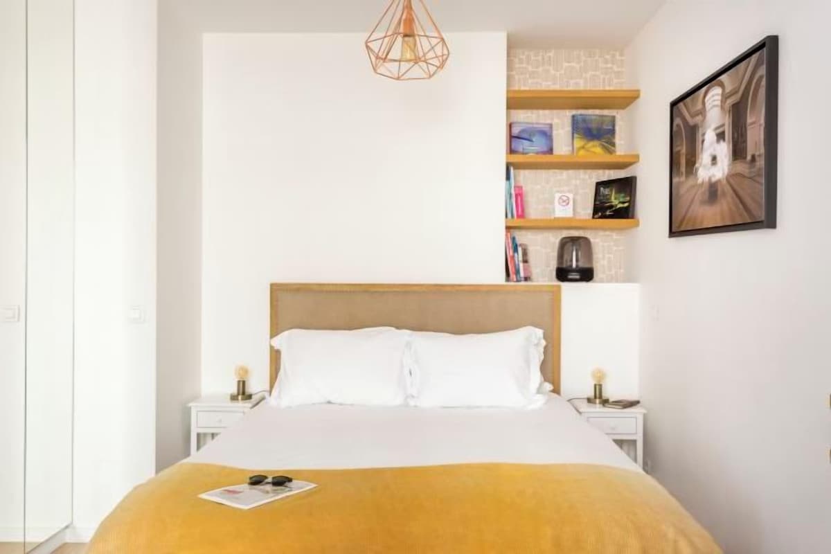 A bedroom with a bed, a bedside table, a nightstand, a bookshelf, and a hanging light fixture.
