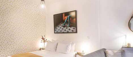 A hotel room with a bed, a gray sofa, a framed picture on the wall, a nightstand with a lamp, and patterned wallpaper.