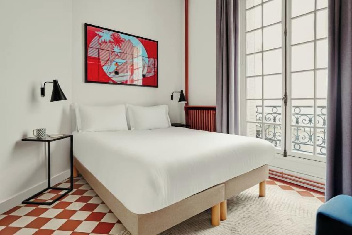 A modern hotel room with a large bed, a red and white checkered floor, a large window with a view, and a piece of abstract art on the wall.