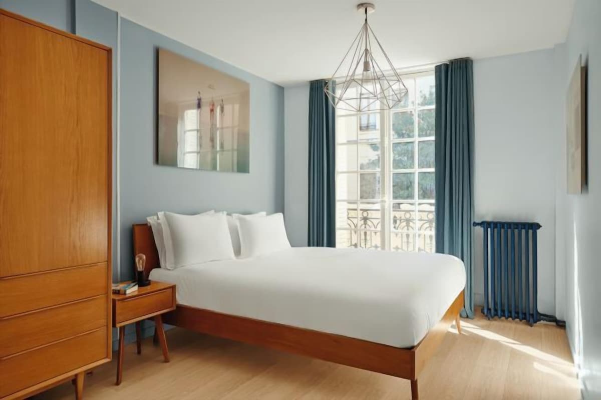 A bedroom with a large bed, wooden wardrobe, a nightstand, a modern chandelier, a window with curtains, and a radiator.