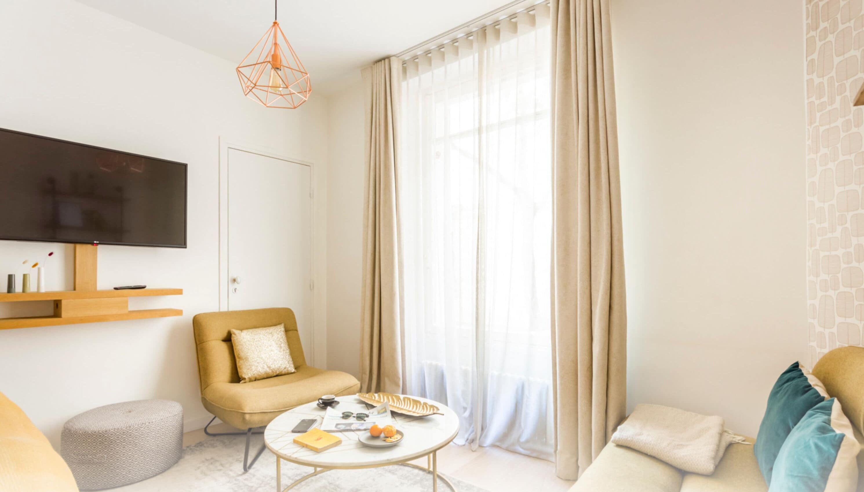 A modern living room with a sofa, a coffee table, a TV mounted on the wall, and a large window with sheer curtains.