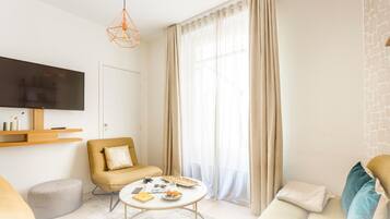 A modern living room with a sofa, a coffee table, a TV mounted on the wall, and a large window with sheer curtains.