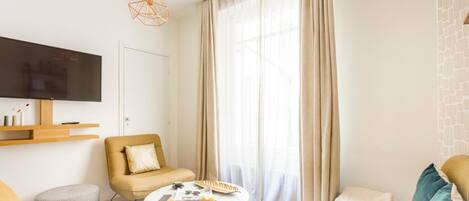 Un salon moderne avec un canapé, une table basse, un téléviseur fixé au mur et une grande fenêtre ornée de rideaux légers.