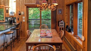 A wooden cabin interior with a dining table, chairs, a kitchen area, and a large window with a view of greenery.