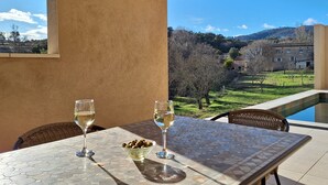 Una mesa con dos copas de vino blanco y un cuenco de aceitunas en un patio empedrado, con vistas a una colina y un edificio.