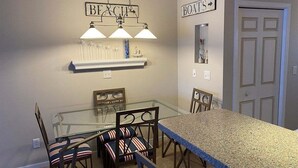 Un comedor con mesa de vidrio, sillas con cojines a rayas, encimera de granito, lámparas colgantes y estanterías en la pared.