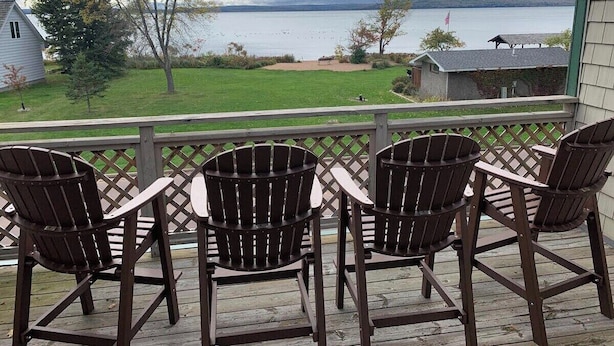 Una terraza con cuatro sillas Adirondack y una pequeña mesa, con vista a un área verde y un cuerpo de agua.