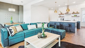 A modern living room with a teal sofa, a wooden coffee table, and a kitchen area with a bar and pendant lights.