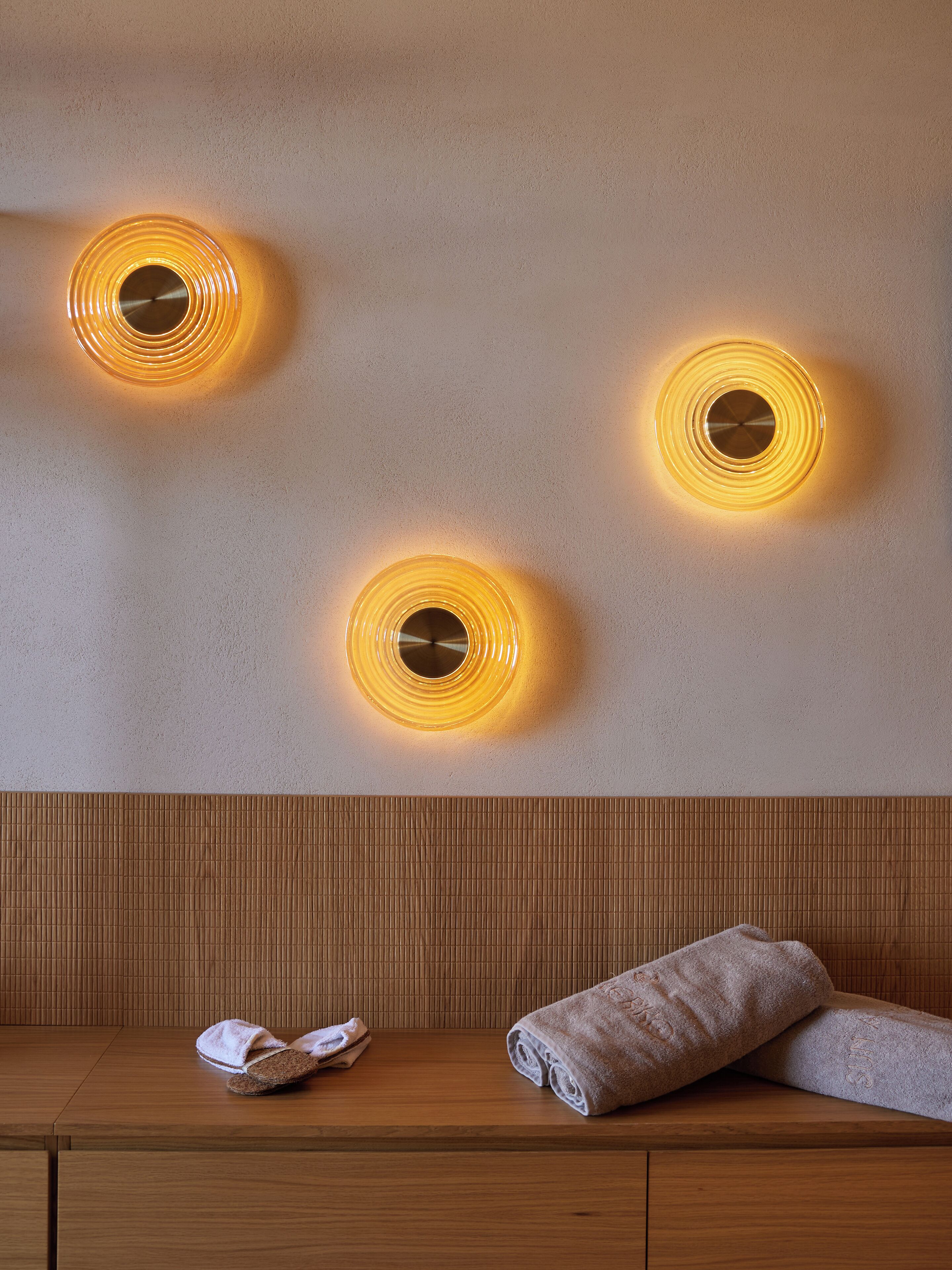A wall with three circular light fixtures and a wooden bench with towels and slippers.