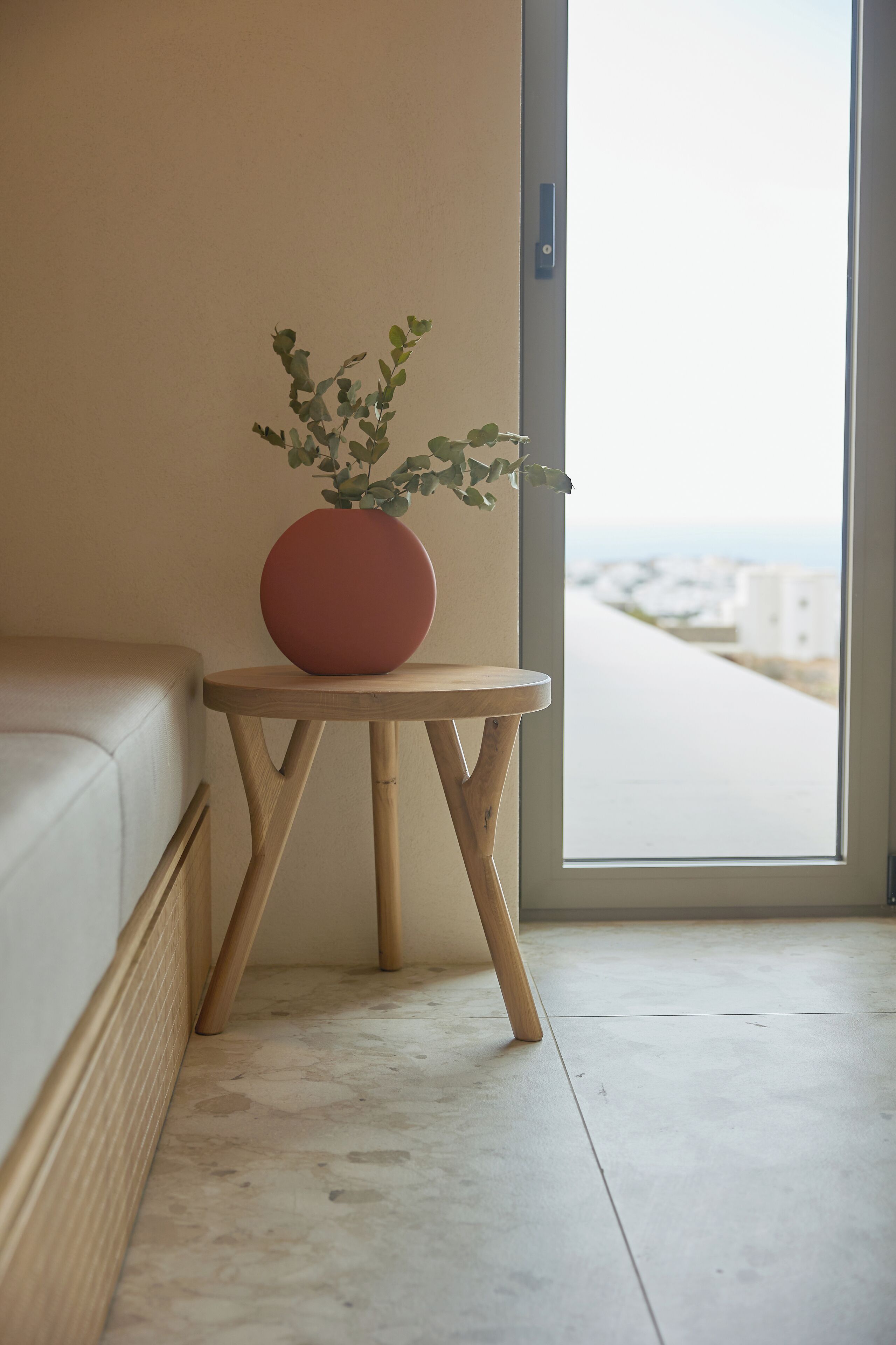 Una habitación con sofá color beige, una mesita de madera con un jarrón rojo y vistas a la costa a través de un ventanal.