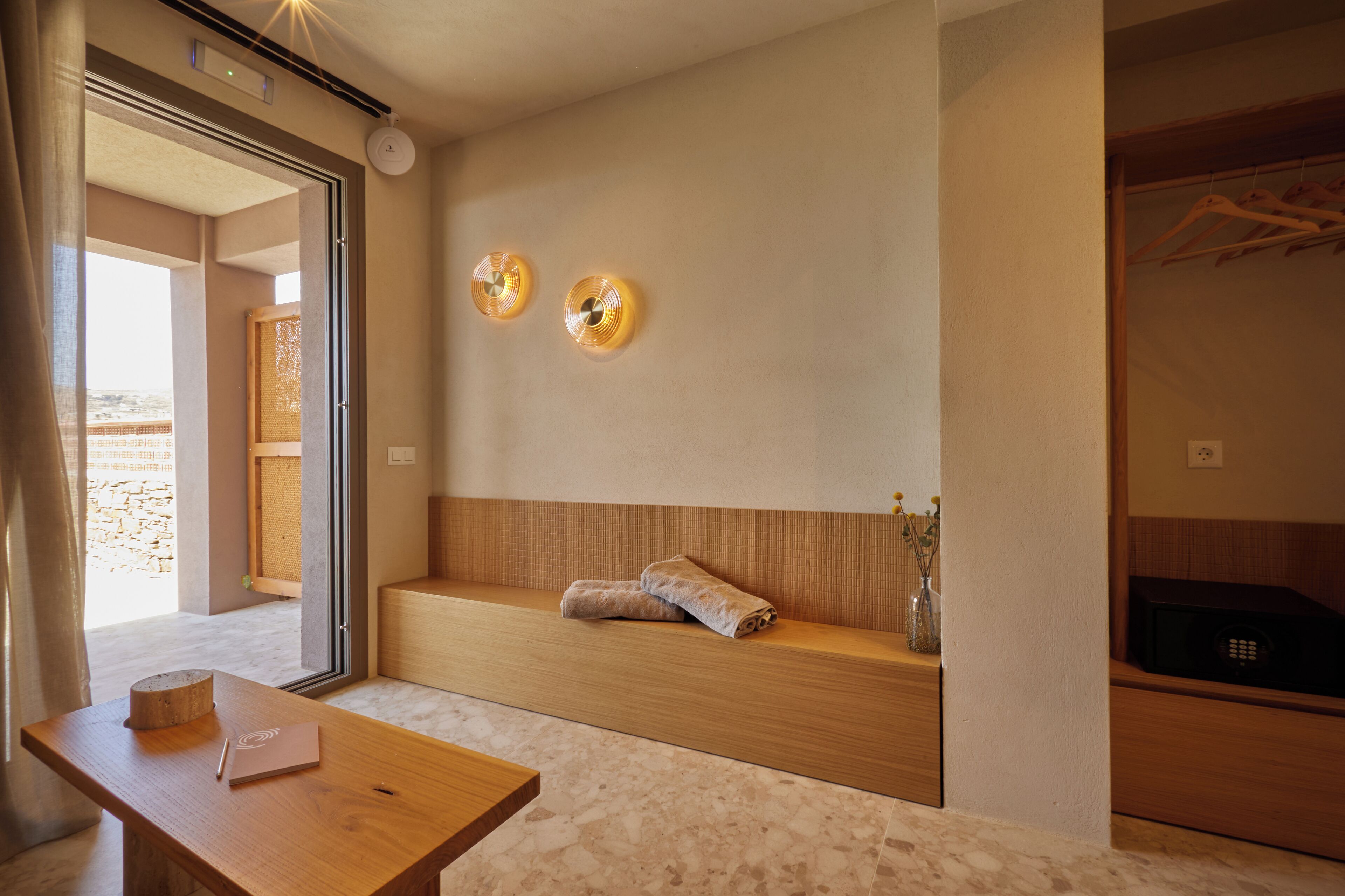 A modern room with a wooden bench, a small table with a notebook and pen, a wall-mounted light fixture, and a view of a stone wall through an open door.