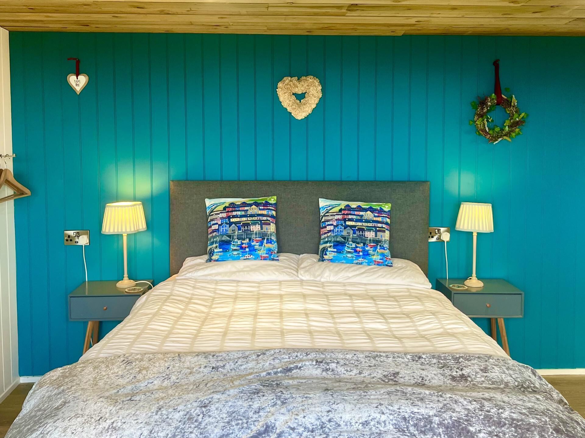 A bedroom with teal walls, a bed with patterned pillows, two bedside lamps, and a heart-shaped decoration on the wall.