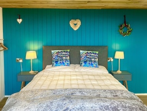 A bedroom with teal walls, a bed with patterned pillows, two bedside lamps, and a heart-shaped decoration on the wall. - The Pod Luxury Accommodation (Aberaeron)