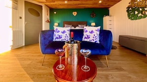 A room with a wooden ceiling, a blue sofa with purple floral cushions, a wooden side table with two wine glasses and a bottle, and a green wall with a heart decoration. - Luxury Pods - Sleeps 2 & Free Parking (Aberaeron)
