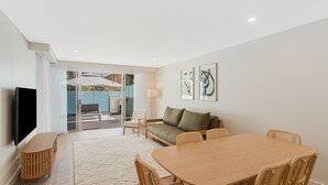 A modern living room with a sofa, dining table, and chairs. There is a TV mounted on the wall and a window with curtains.