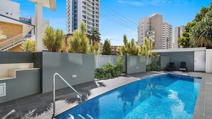 A modern swimming pool with a metal ladder, surrounded by a grey wall and lush greenery.