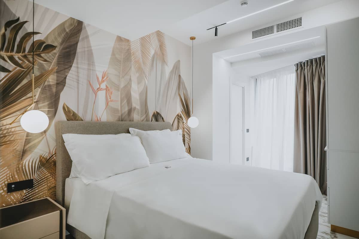 A modern hotel room with a large bed, a nightstand, a wall with tropical leaf patterns, and a door with curtains.