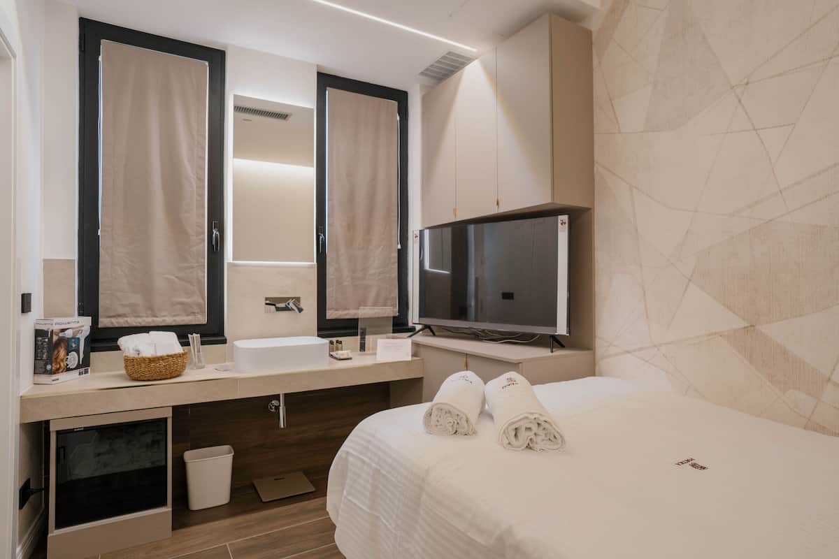 A hotel room with a bed, two mirrors, a television, a sink, and a small trash bin.
