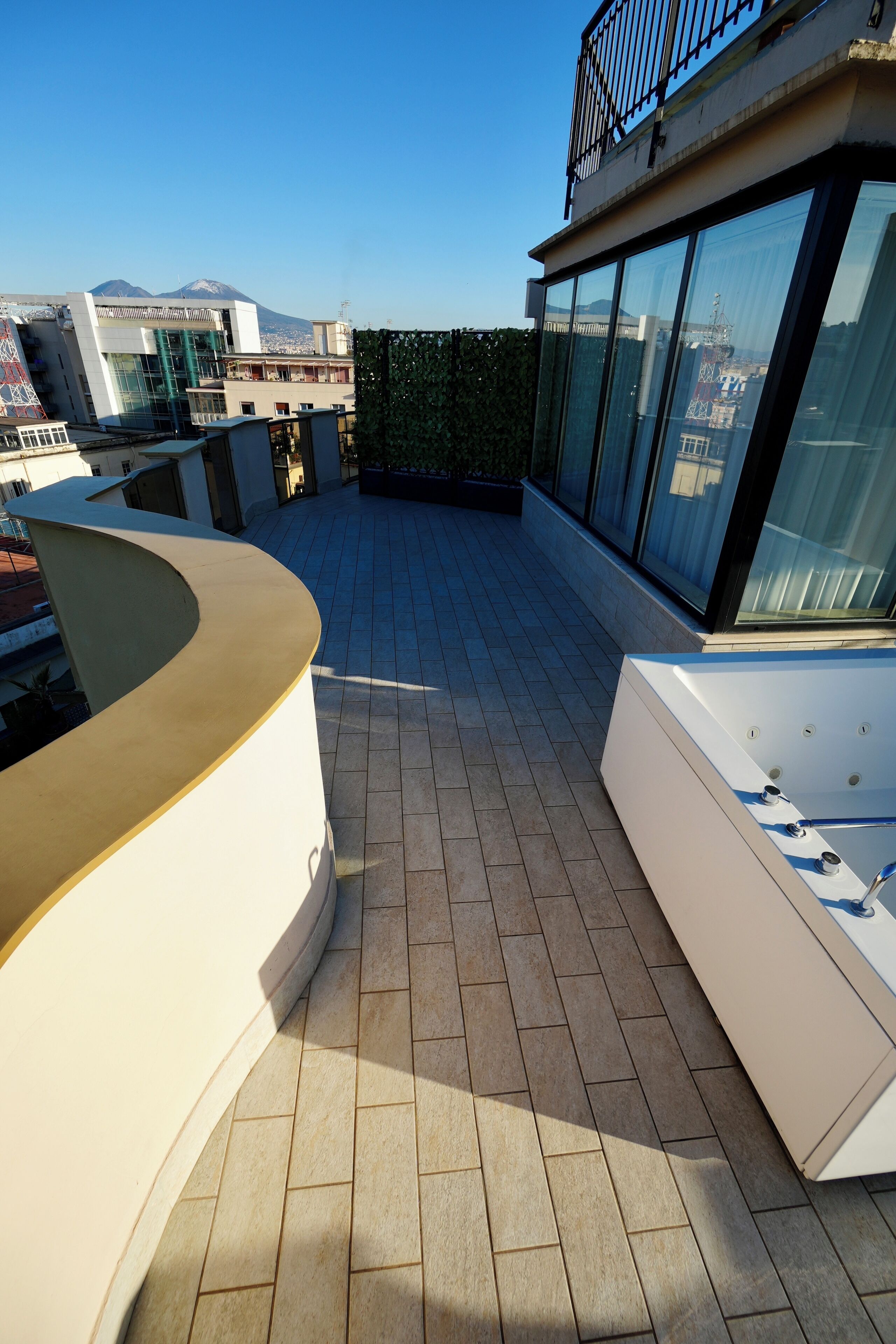 A rooftop terrace with a curved white bench, a spa tub, and a glass balustrade.