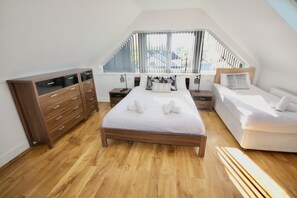 A bedroom with a wooden floor, a large bed with white bedding, a wooden dresser, and a window with blinds.