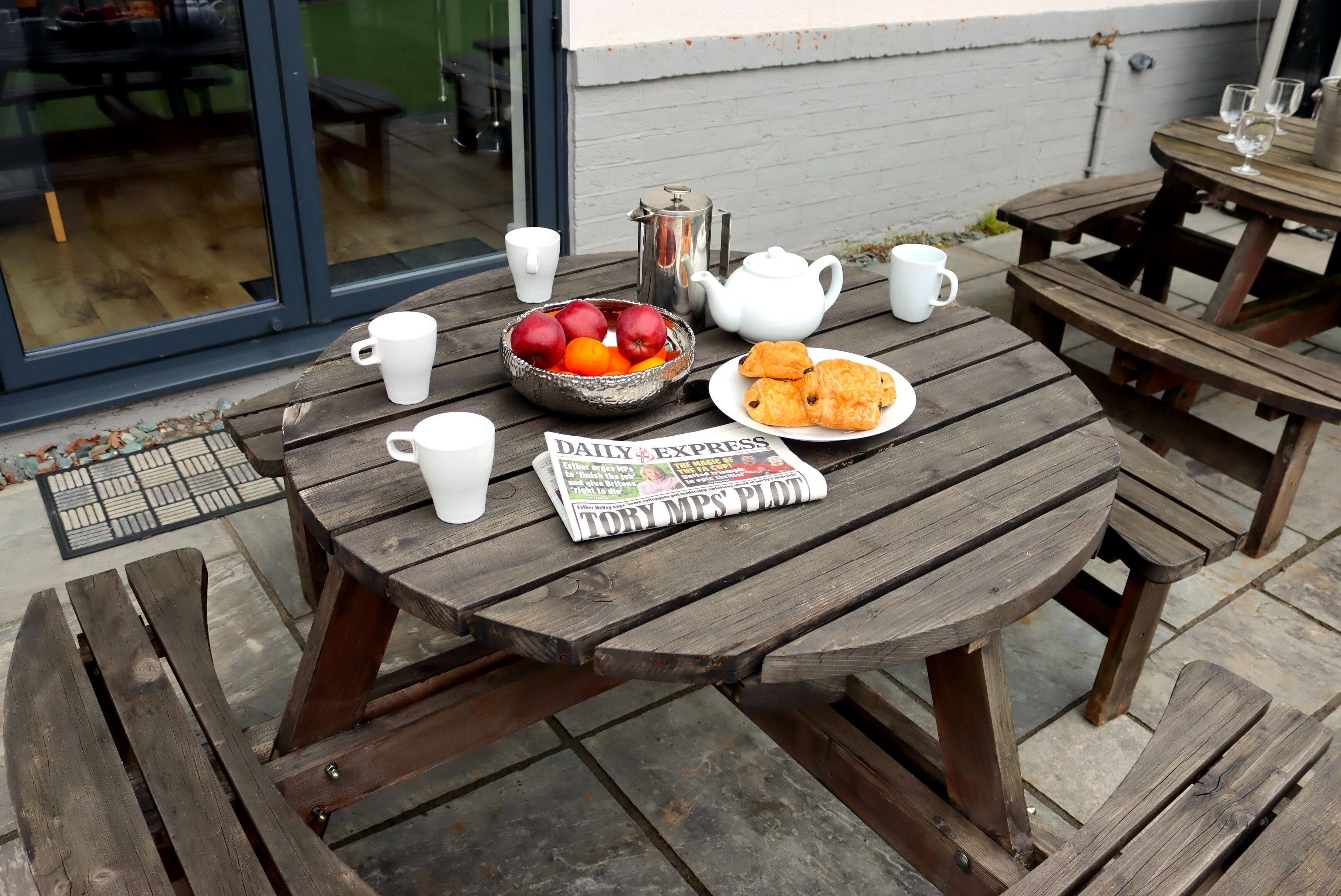 A wooden picnic table with a newspaper, a bowl of fruit, a teapot, and a plate of pastries.