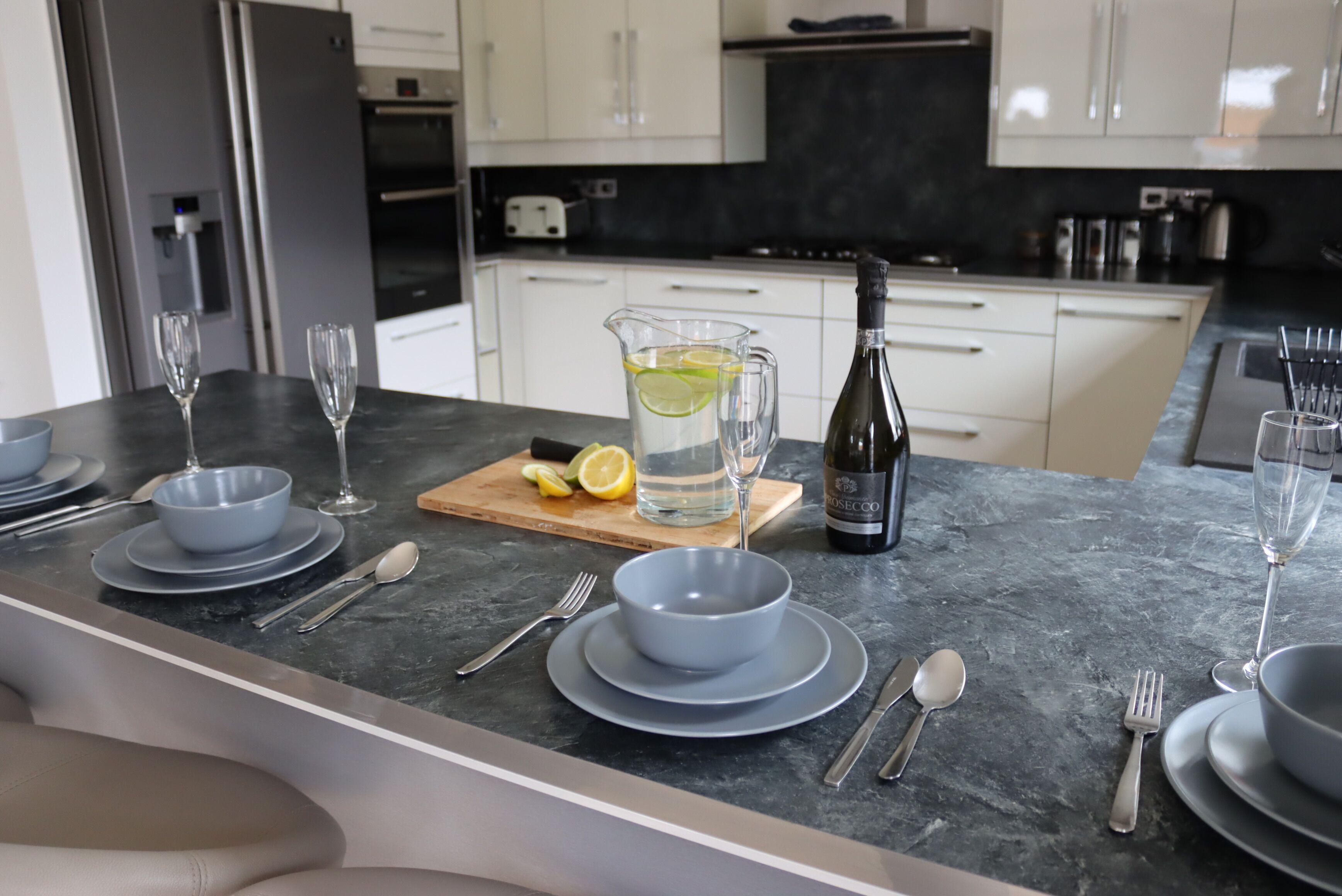 A modern kitchen with white cabinets, a marble countertop, and a dining area set with plates, bowls, cutlery, and a pitcher of water with lemon slices.