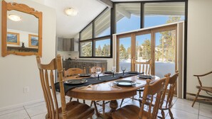 A dining area with a round wooden table set for six, surrounded by wooden chairs. A large mirror hangs on the wall, and there is a view of a snowy landscape through the glass doors. - 45 Idlewild New 2 bedroom private home private hot tub, walk to downtown (Winter Park)