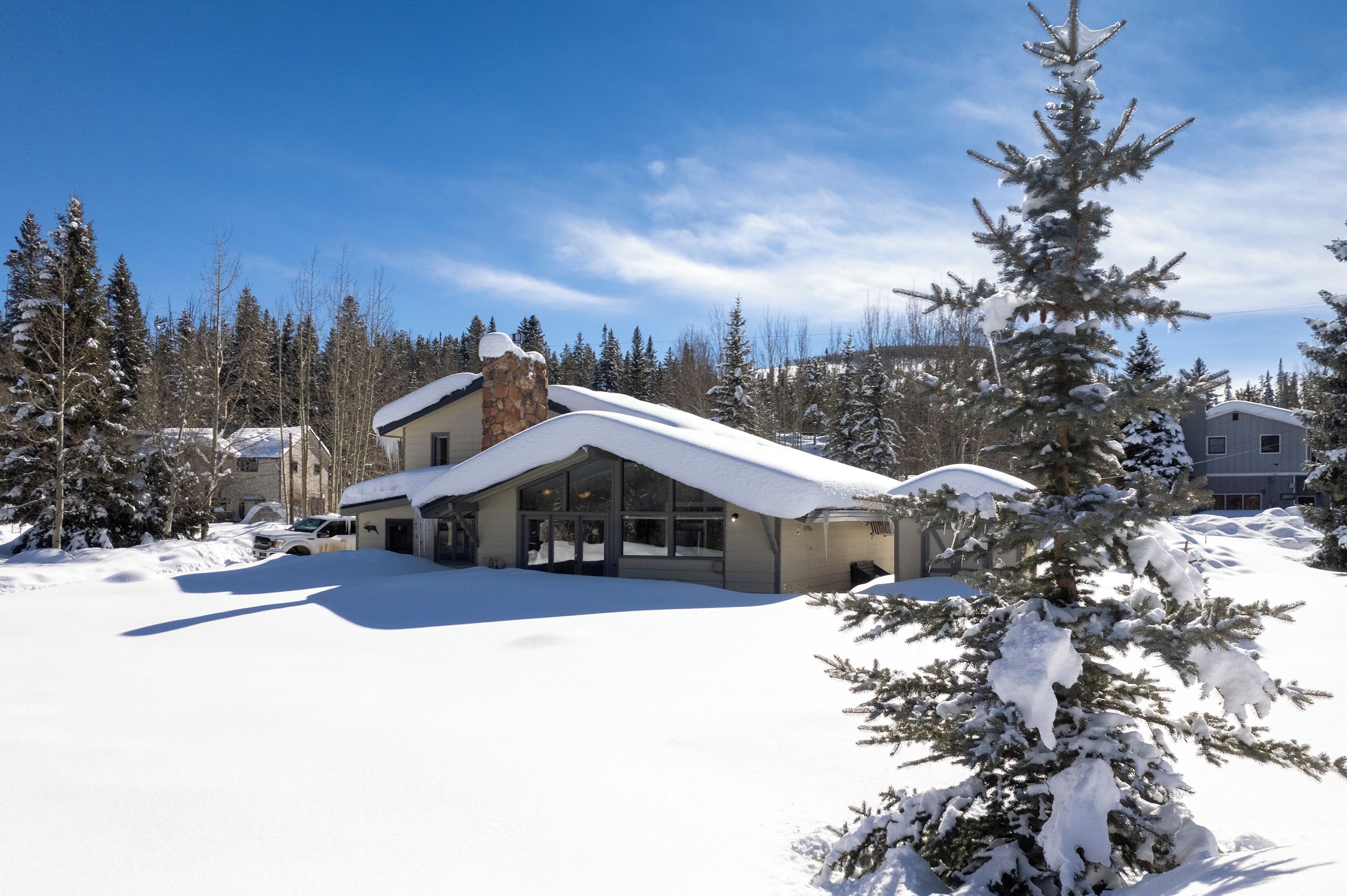 Ein schneebedecktes Haus mit einem Steinkamin, umgeben von mit Schnee beladenen Bäumen und einem klaren, blauen Himmel.