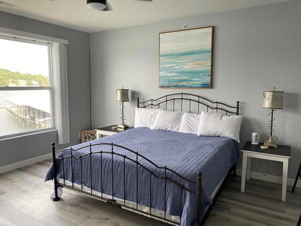 Quarto com cama, duas mesas de cabeceira, janela com vista para um cais e um quadro na parede.