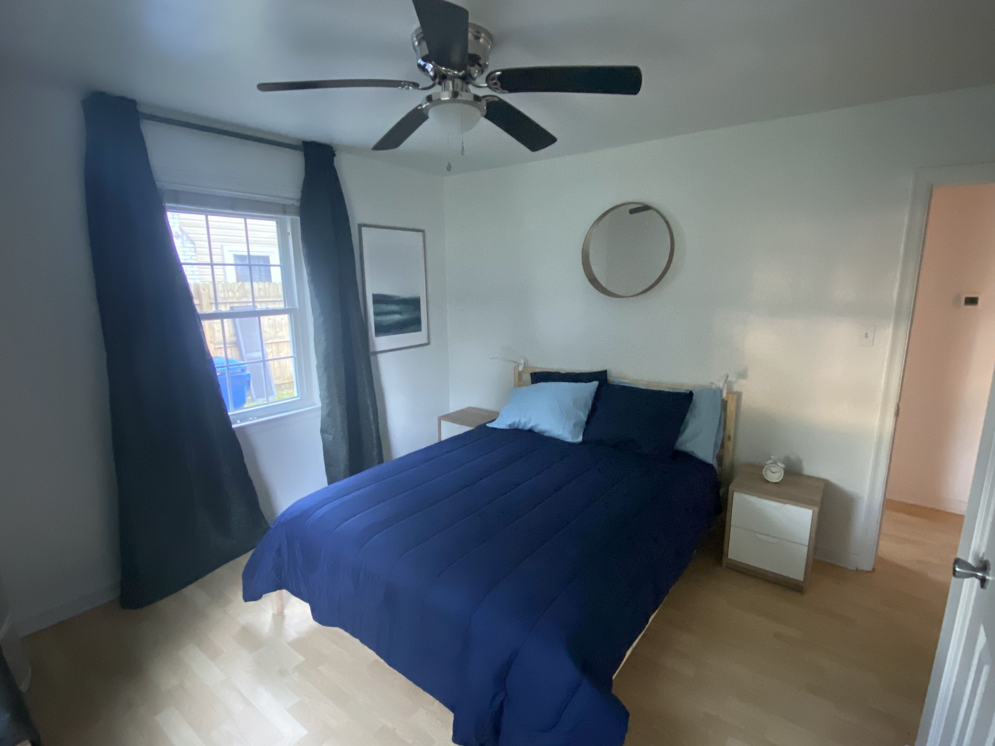 A bedroom with a bed, a ceiling fan, a bedside table, a clock, and a window with curtains.