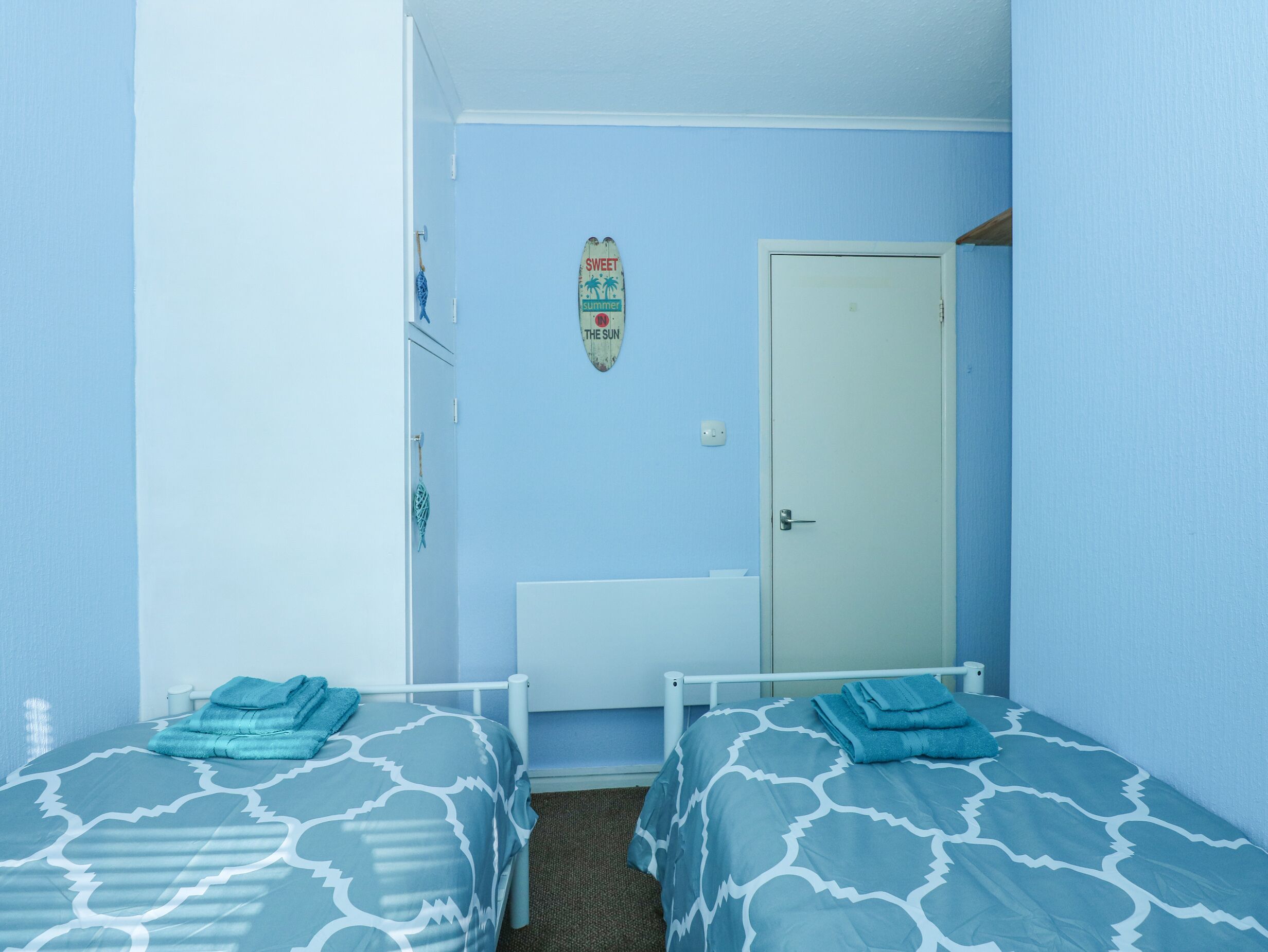 Een kleine hotelkamer met één slaapkamer, met twee bedden, elk met een blauw beddegoed met patroon. Er is een deur met een bordje erboven, een klein plankje met spullen, en een lichtschakelaar aan de muur.