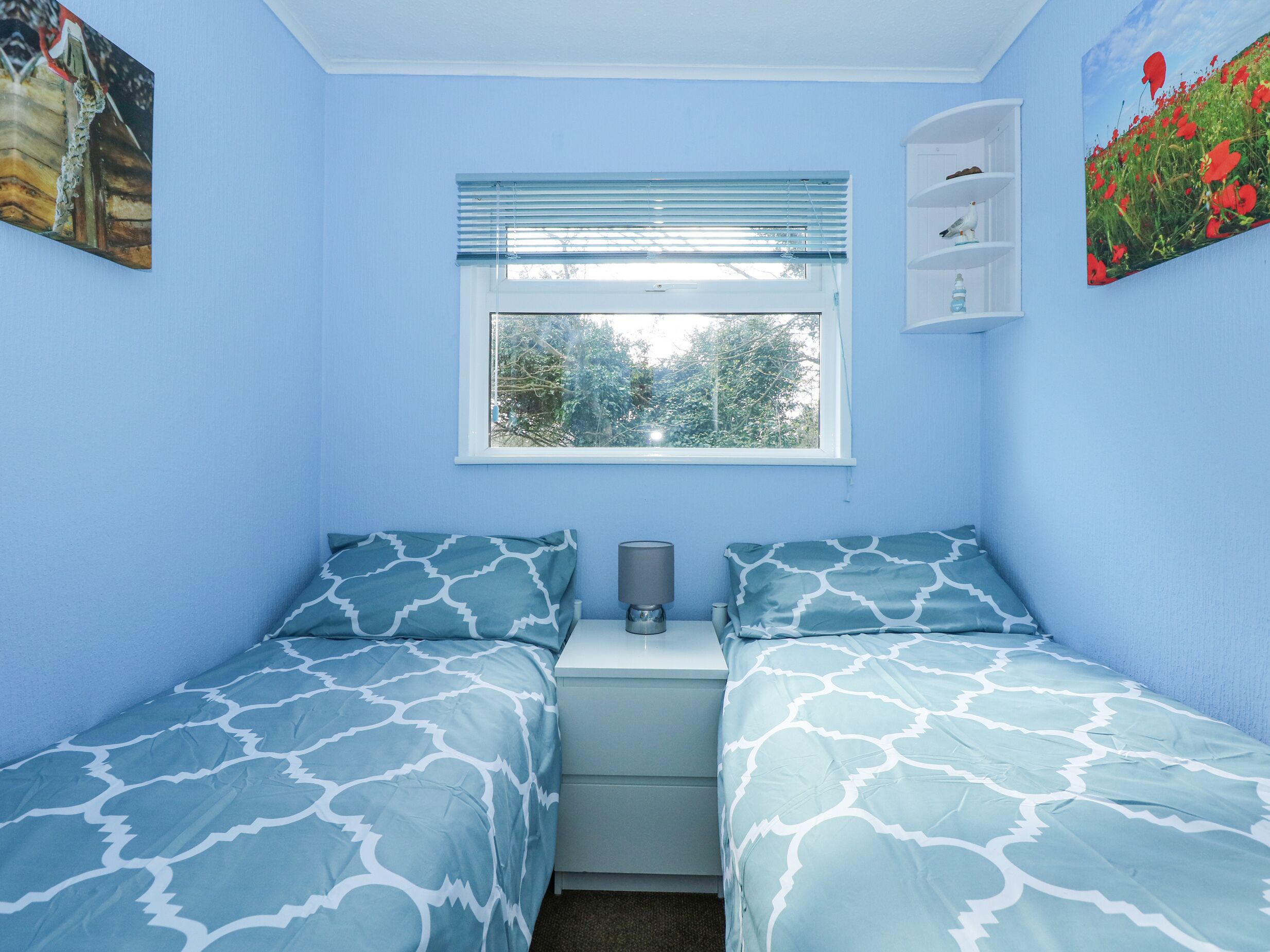 Un dormitorio con dos camas, una ventana con persianas, una mesita de noche con lámpara y una estantería con objetos decorativos.