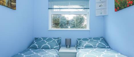 Un dormitorio con dos camas, una ventana con persianas, una mesita de noche con lámpara y una estantería con objetos decorativos.