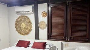 Ein Schlafzimmer mit einem Bett, zwei Kissen, einem Sessel, einem roten Teppich, einer Wand-Klimaanlage und zwei Holztüren.