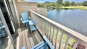 Une terrasse en bois avec des chaises Adirondack bleues et une table, donnant sur un lac paisible et un terrain de golf.
