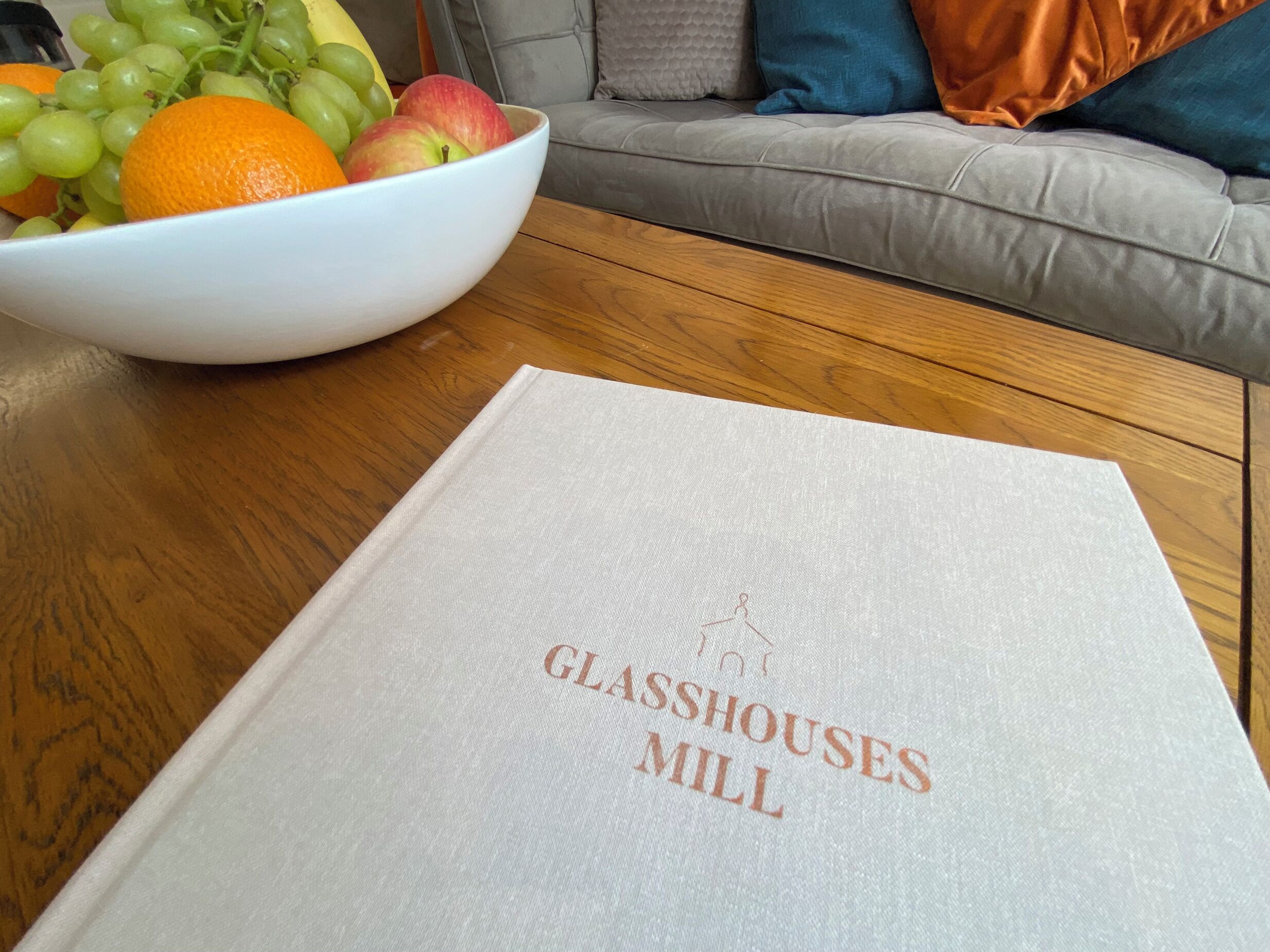 Eine Schale mit Obst auf einem Holztisch, vor ihr ein Buch mit dem Titel 'Glasshouses Mill'.