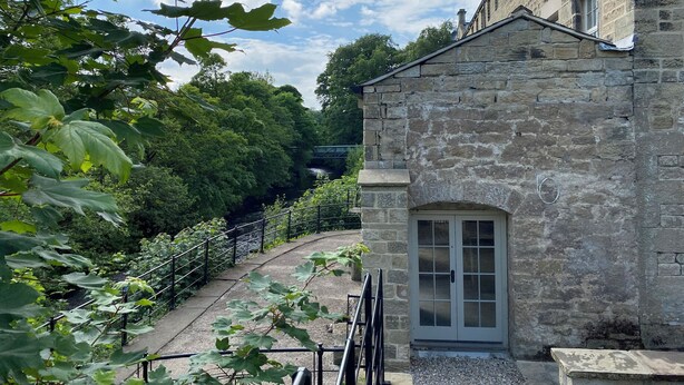 A stone building with a gated entrance, a pathway leading to a bridge, and lush greenery in the background. - Millnook (Harrogate)