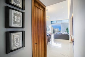 A hallway with framed artwork on the wall, a wooden door, and a view into a modern living area with a dining table and chairs.