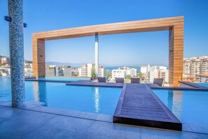 Ein Pool mit Holzterrasse und Liegestühlen, mit Blick auf eine Stadtlandschaft und das Meer.