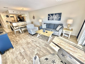 A living room with a striped armchair, a grey sofa, a coffee table, and a blue dining table. There is a kitchen in the background with a chandelier.